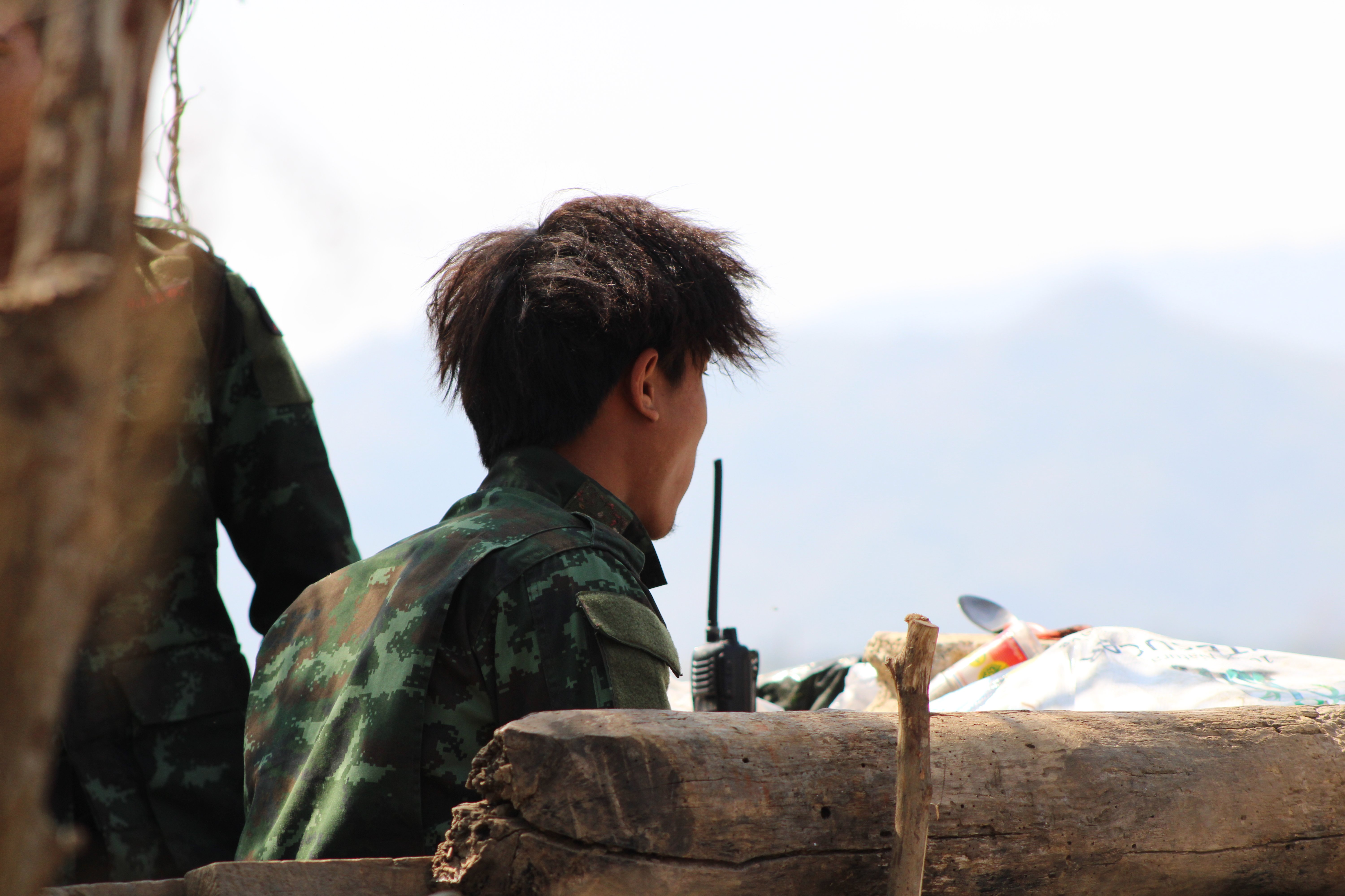 Soldier in camouflage uniform using a radio, seated on a wooden structure with a mountainous background.