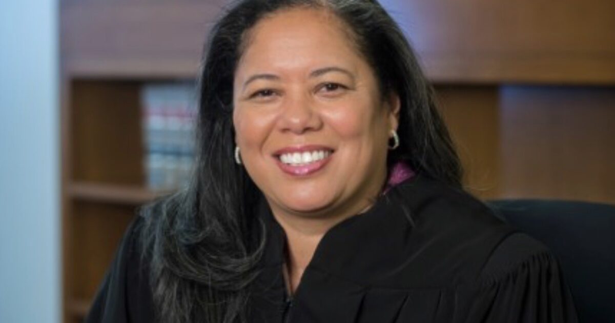 Smiling female judge in a black robe seated in an office with law books in the background, representing authority and professionalism in the legal field.