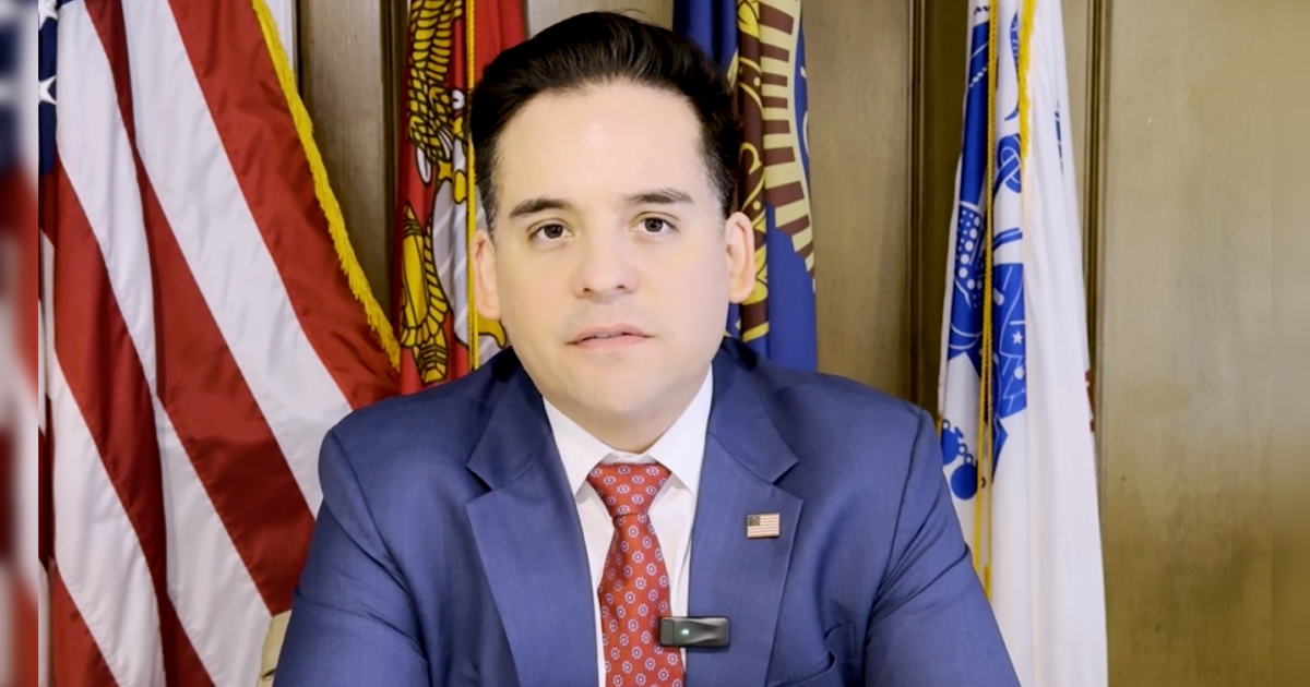 Man in a blue suit with a red tie speaking in front of multiple flags, conveying a message or announcement.
