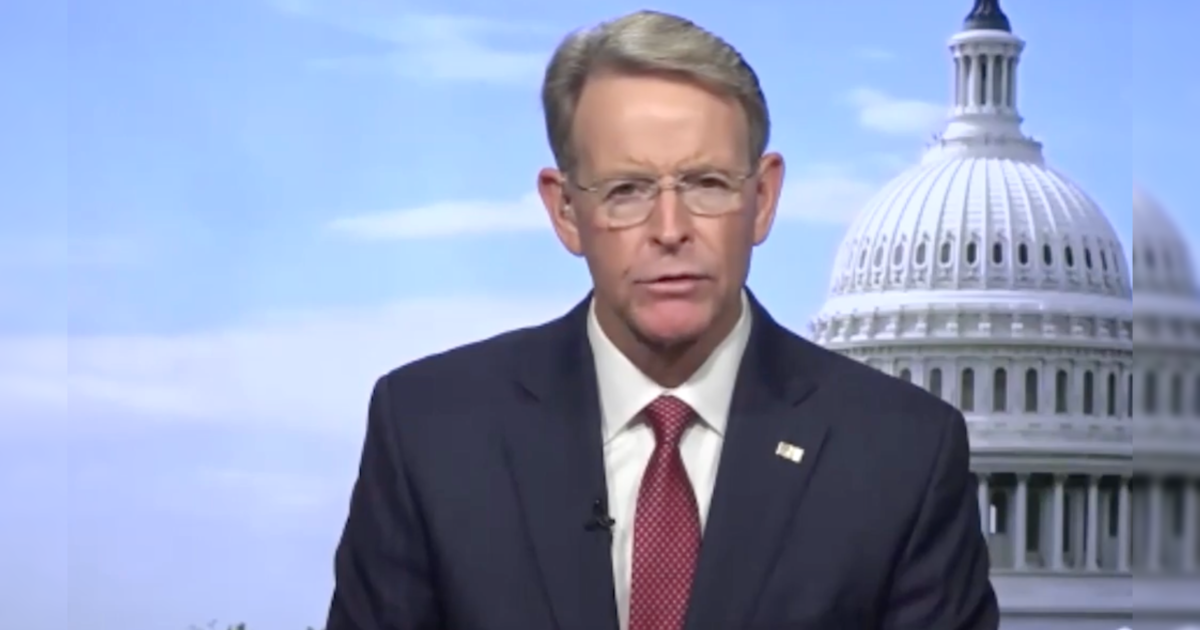 Man in a suit speaking in front of the U.S. Capitol building, discussing political issues on a news broadcast.