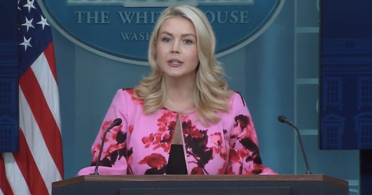 Press secretary addressing the media at the White House briefing room, wearing a floral pink blazer, with American flag in the background.