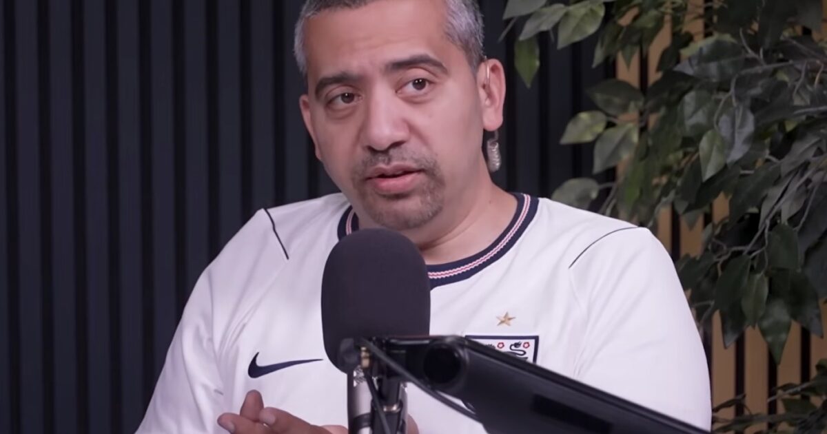 A man in a white sports jersey speaks into a microphone during a podcast, surrounded by greenery and a modern studio backdrop.