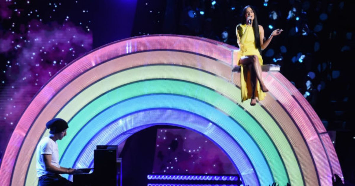 A singer in a yellow dress performs on a colorful rainbow stage while a pianist plays, set against a starry background.