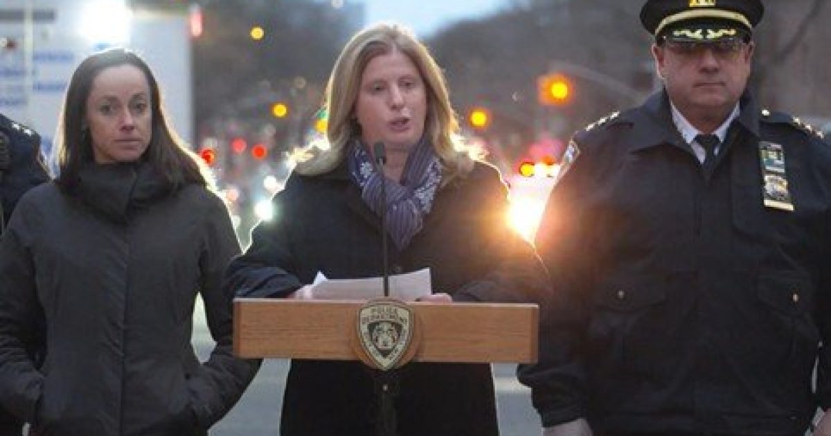 Press conference featuring officials addressing the media, with a female speaker at a podium, flanked by two colleagues in formal attire, set against a city backdrop.